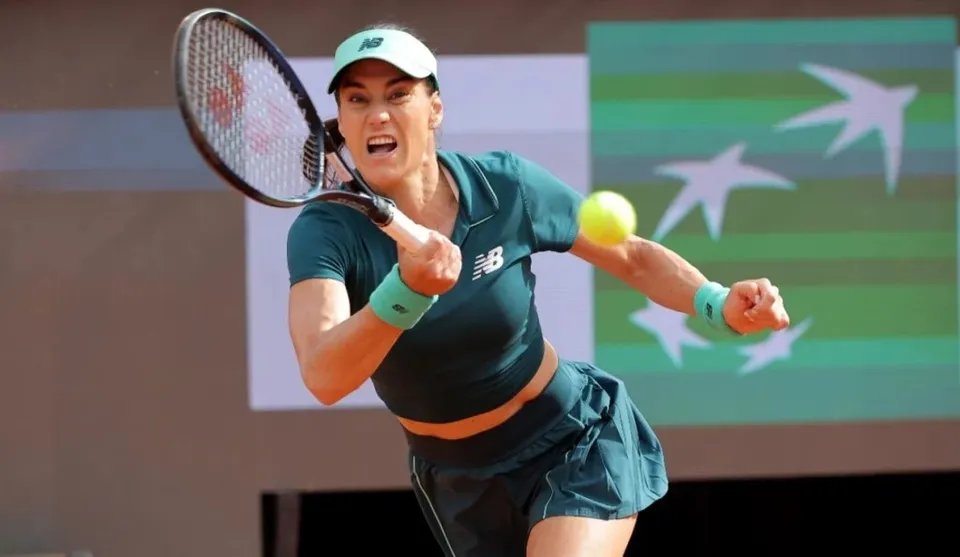 O femeie cu o cascadă de păr scurt și șapcă albă lovește o minge galbenă cu o rachetă de tenis, purtând o echipă de culoare verde închis.