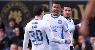 Trei jucători de fotbal în echipamente albe cu detalii albastre sărbătoresc pe un teren de sport.