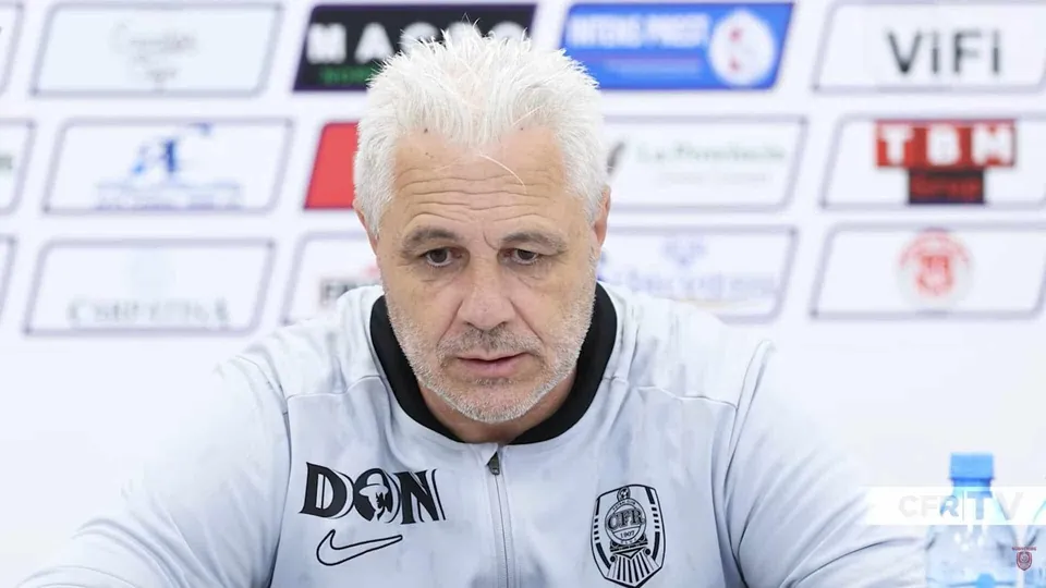 Un bărbat cu părul alb și scurt stă în fața unui fundal alb cu diverse logo-uri colorate.