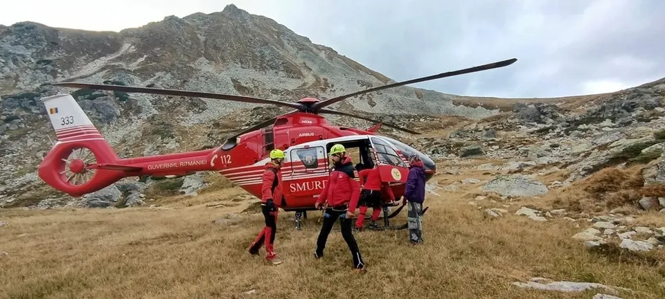 Un elicopter roșu cu inscripția SMURD este aterizat pe un munte, înconjurat de patru persoane purtând haine colorate și casci de protecție.