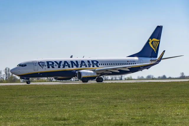 Un avion de pasageri alb cu însemnele Ryanair taxiind pe o pistă de aterizare lângă un câmp de iarbă verde sub un cer senin.