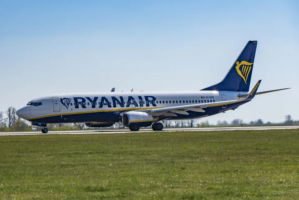 Un avion de pasageri alb cu însemnele Ryanair taxiind pe o pistă de aterizare lângă un câmp de iarbă verde sub un cer senin.