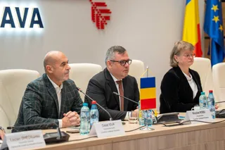 Trei persoane stau la o masă de conferință cu microfoane, sticle de apă și plăcuțe de identificare în fața unor steaguri.