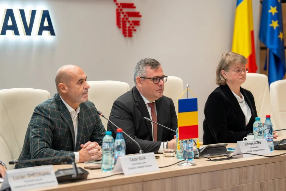 Trei persoane stau la o masă de conferință cu microfoane, sticle de apă și plăcuțe de identificare în fața unor steaguri.