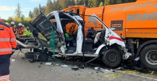 Un microbuz alb cu caroseria puternic distrusă stă pe o stradă lângă un vehicul utilitar portocaliu și persoane purtând echipamente de protecție reflectorizante.