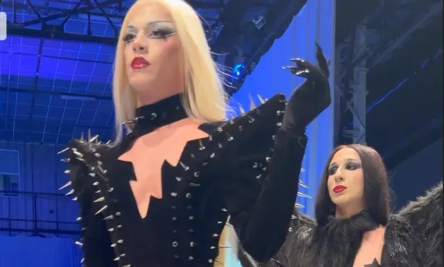 O persoană cu păr blond lung și machiaj dramatic poartă o vestimentație neagră cu țepi metalice și mănuși lungi pe un fundal albastru.