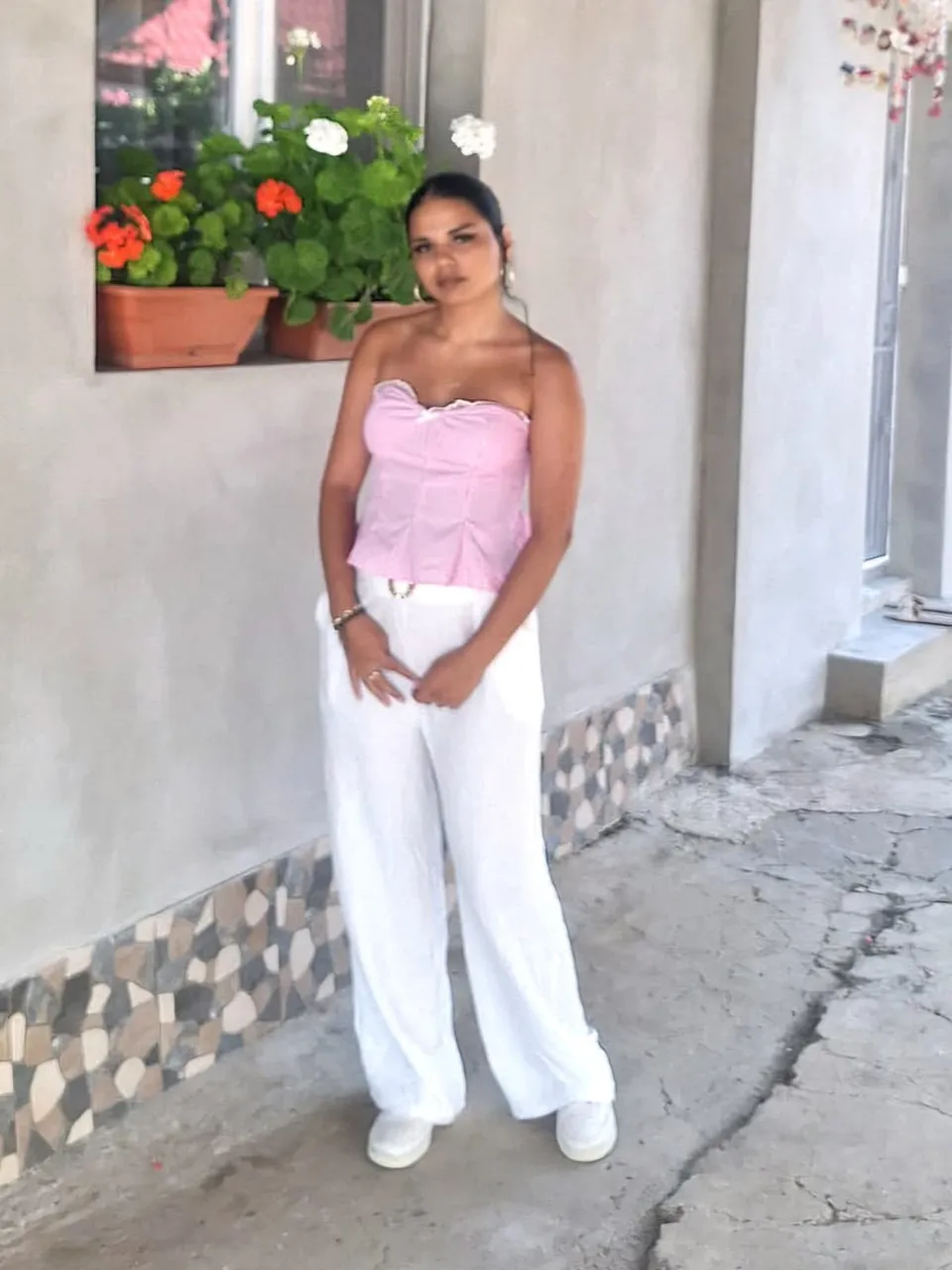 O tânără poartă un bustier roz și pantaloni albi largi, stând pe un trotuar lângă un perete cu ghivece cu flori.