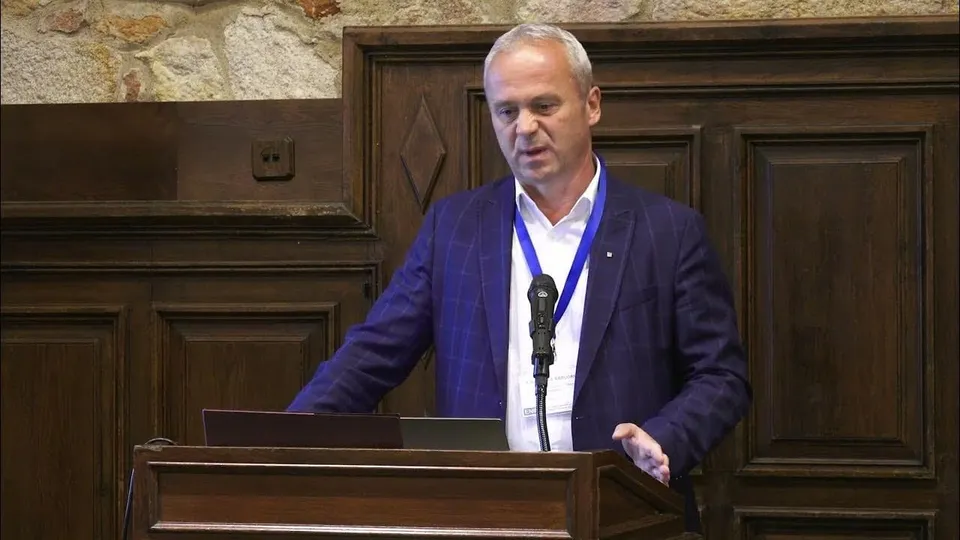Un bărbat cu părul scurt și cămașă albă, purtând un sacou albastru și un cordon albastru, stă în spatele unui pupitru de lemn.
