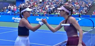 Două femei cu viziere și haine de sport se strâng în mâini pe un teren de tenis de culoare albastră în fața unui public.