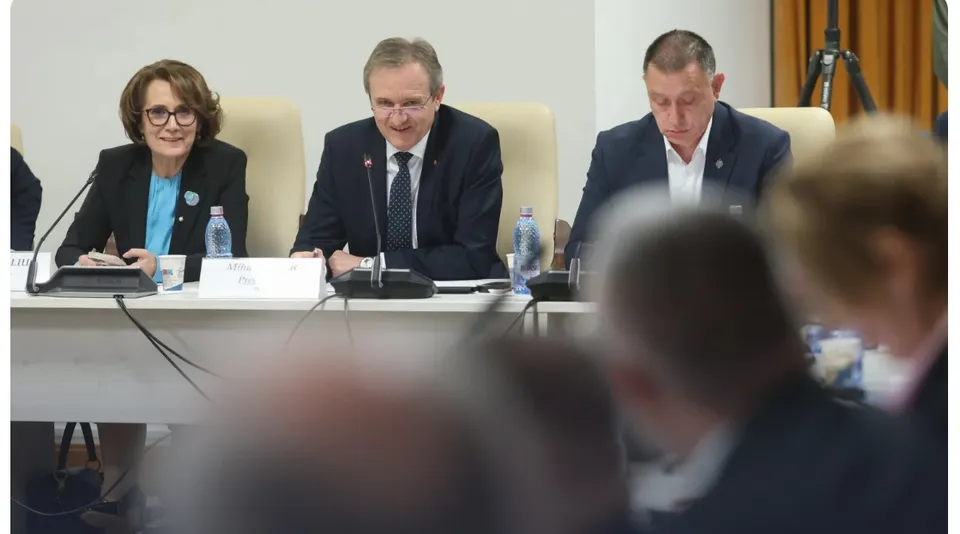 Trei persoane stau la o masă lungă de conferință, purtând haine office, în fața unor microfoane și sticle de apă pe o suprafață albă.