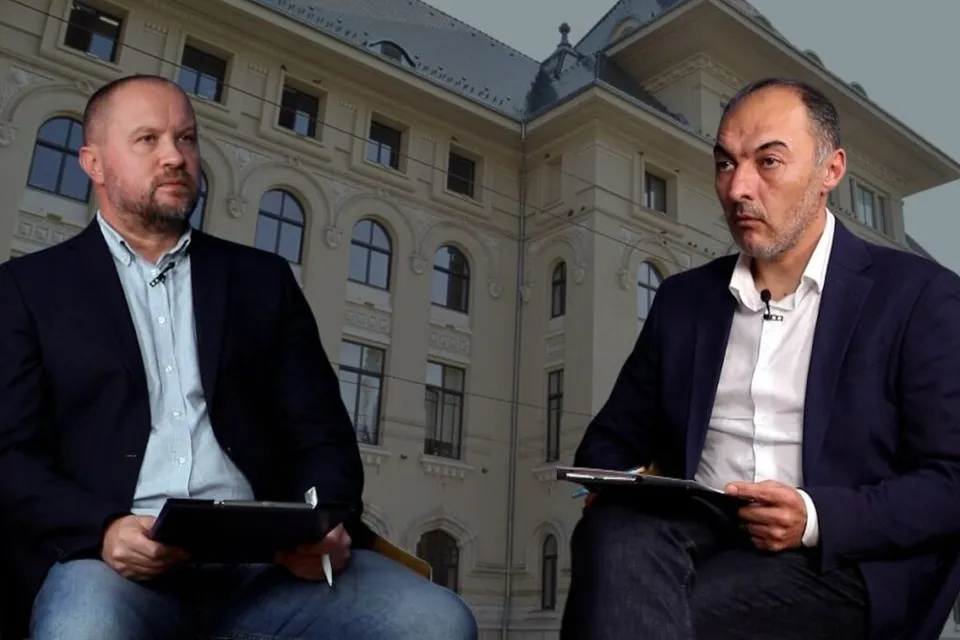 Doi bărbați cu sacou și cămașă stau așezați în fața unei clădiri înalte cu ferestre multiple.