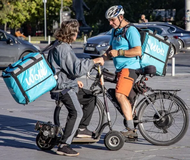 Un bărbat cu cască și un sac albastru de livrări stă lângă un alt bărbat pe un scuter electric și o bicicletă.