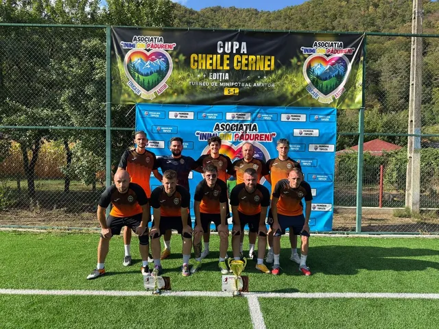 Fotbaliiști în tricouri negre și portocalii posedă lângă două trofee, în fața unor bannere cu inscripții, pe un teren sintetic.