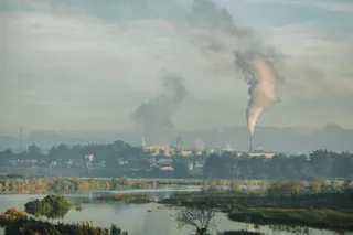 Fumul alb se ridică de la un coș de fum de deasupra unui complex industrial situat lângă un corp de apă.