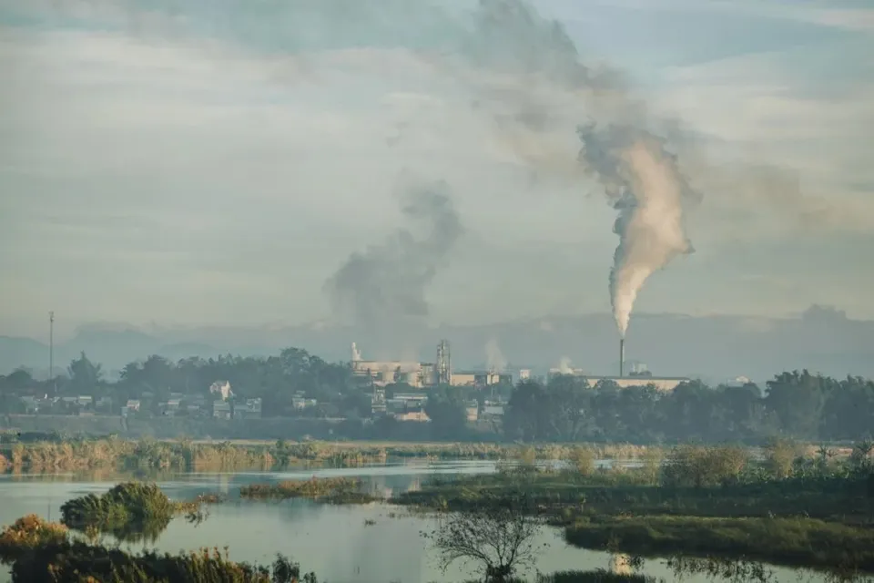 Fumul alb se ridică de la un coș de fum de deasupra unui complex industrial situat lângă un corp de apă.