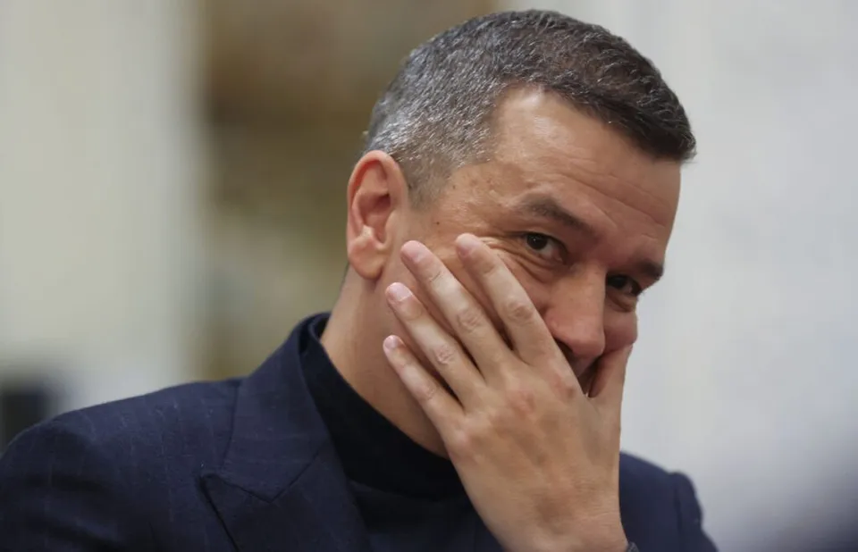 Un bărbat cu părul scurt și scurtat pe lateral poartă un sacou închis peste un tricou negru, având mâna pe față.