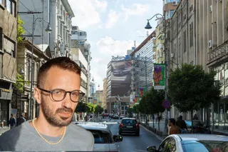 Un bărbat cu ochelari și un colier de perle este în prim-plan, în fața unei străzi urbane cu mașini și clădiri înalte.