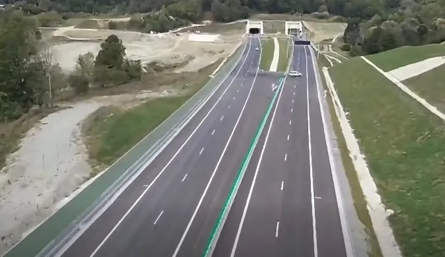 O autostradă cu mai multe benzi de rulare traversează un peisaj cu dealuri verzi și zone de teren excavat sub un tunel.
