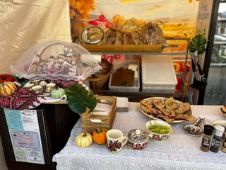 Diverse produse alimentare, fructe, dovleci și ustensile de ceramică sunt expuse pe o masă acoperită cu o față de masă albă sub un prelat.