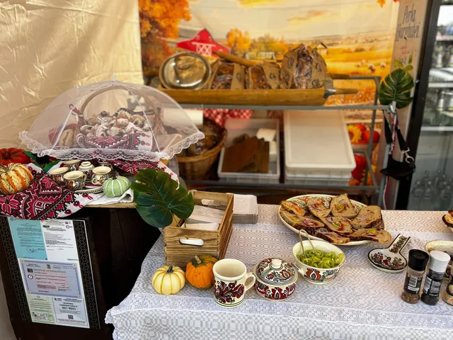 Diverse produse alimentare, fructe, dovleci și ustensile de ceramică sunt expuse pe o masă acoperită cu o față de masă albă sub un prelat.