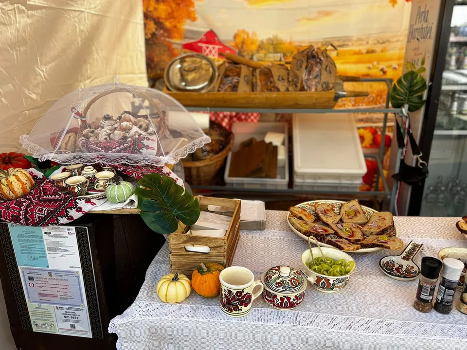 Diverse produse alimentare, fructe, dovleci și ustensile de ceramică sunt expuse pe o masă acoperită cu o față de masă albă sub un prelat.