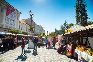 Oamenii se plimbă pe un drum pavat între tarabe de vânzare cu produse și clădiri sub un cer senin.