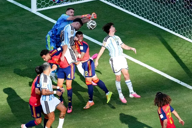 Câțiva jucători de fotbal în echipamente albastre, albe și roșii se află pe un teren verde lângă poarta de fotbal, încercând să atingă un balon.