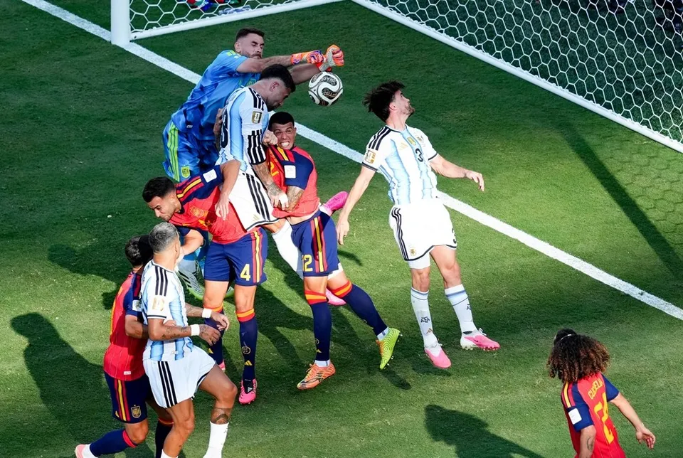 Câțiva jucători de fotbal în echipamente albastre, albe și roșii se află pe un teren verde lângă poarta de fotbal, încercând să atingă un balon.