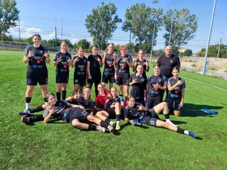 Un grup de femei și fete în echipamente sportive negre cu accente albastre pozează pe un teren de fotbal cu iarbă verde sub cerul senin.