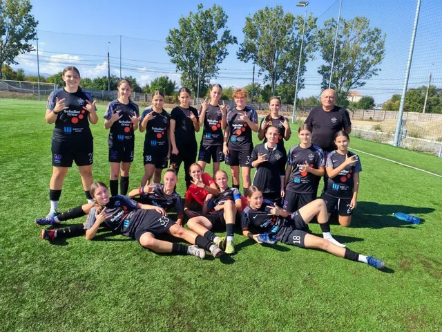 Un grup de femei și fete în echipamente sportive negre cu accente albastre pozează pe un teren de fotbal cu iarbă verde sub cerul senin.