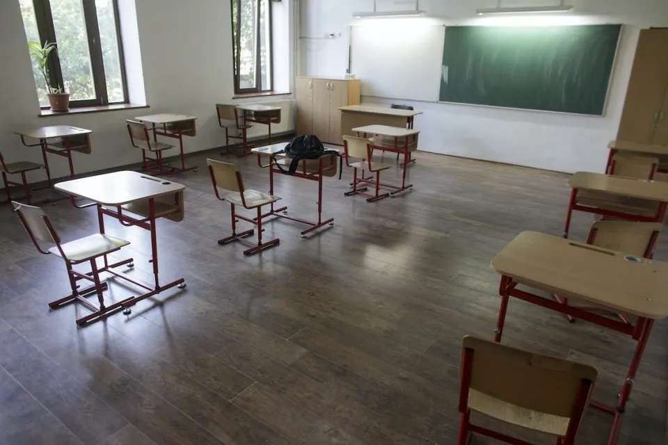 O sală de clasă cu mai multe birouri și scaune de lemn dispuse pe un podea de lemn în fața unui panou verde.