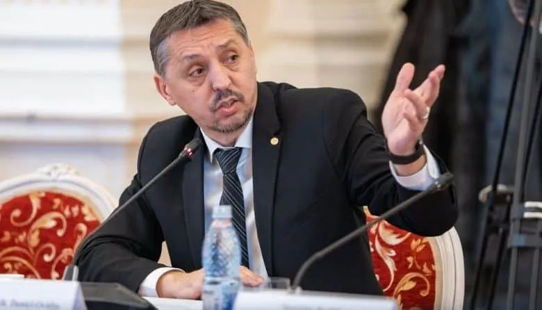 Un bărbat cu mustață, purtând costum negru și cravată închisă, stă la o masă și face un gest cu mâna dreaptă ridicată.