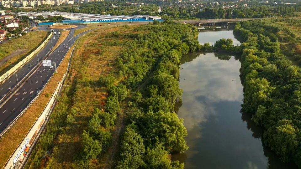 O vedere aeriană prezintă un râu cu vegetație densă pe maluri, alături de o autostradă cu mașini și câteva clădiri în fundal.