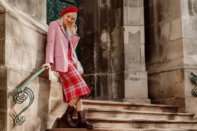 O femeie cu beretă roșie, sacou roz și fustă midi în model tartan pătează pe scări, purtând cizme maro de culoare închisă.