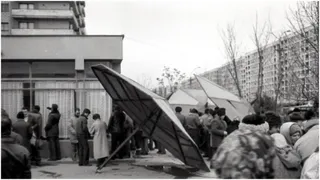 O grupă de oameni stă lângă o structură metalică de formă triunghiulară în fața unui bloc de apartamente.