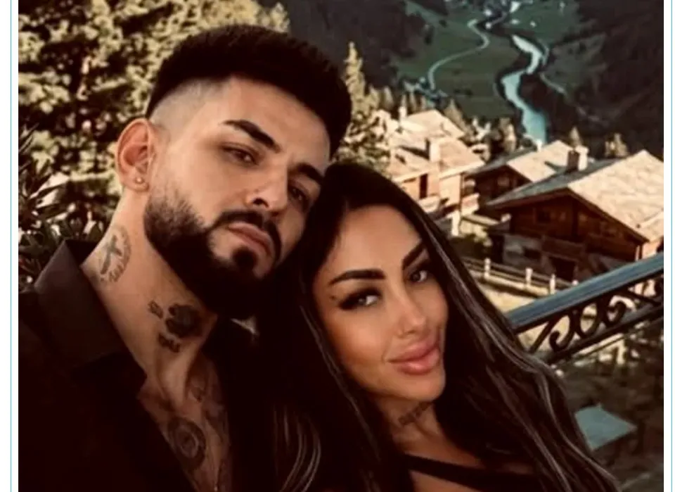 Un bărbat cu barbă și tatuaje pe gât stă lângă o femeie cu părul lung și deschis, în fața unui peisaj montan cu case și un râu.