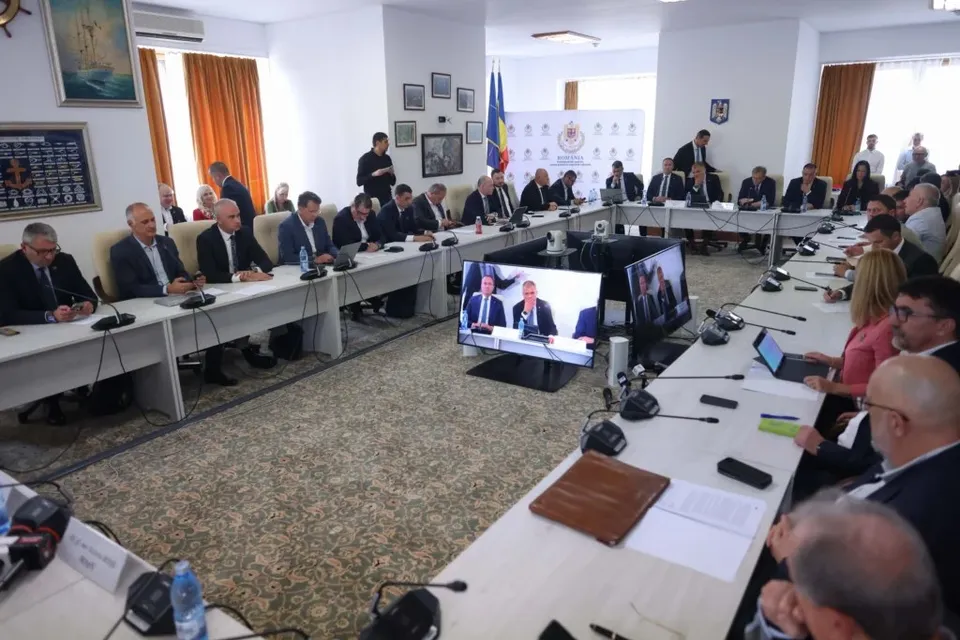 Oamenii stau la mese lungi albe în fața unor ecrane, într-o încăpere cu pereți albi și draperii portocalii.