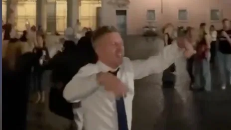 Un bărbat într-o cămașă albă și cravată este surprins în mișcare, cu brațele ridicate, într-un spațiu deschis cu persoane în fundal.