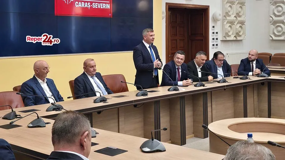 Șase bărbați în costume purtând cămăși și cravate stau așezați sau stau în picioare la o masă lungă de conferințe cu microfoane.