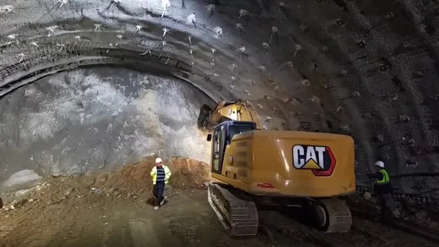 O excavatoare galbenă cu marcajul CAT stă într-un tunel întunecat lângă un lucrător cu cască albă și vestă reflectorizantă pe un drum de pământ.