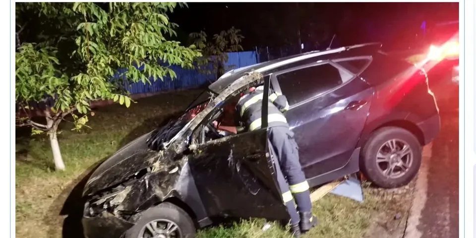 Un salvator în echipament de protecție lucra lângă un autoturism de culoare închisă cu partea frontală zdrobită, aflat lângă un copac noaptea.