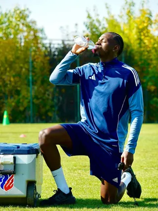 Un bărbat cu haine sportive albastre stă în genunchi pe iarbă și bea apă dintr-o sticlă transparentă.