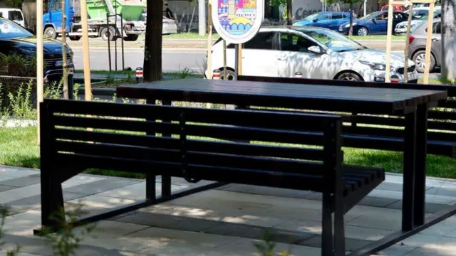 O bancă neagră de lemn este poziționată în prim-plan, în fața unei mese de exterior și a unor mașini parcate pe un drum.