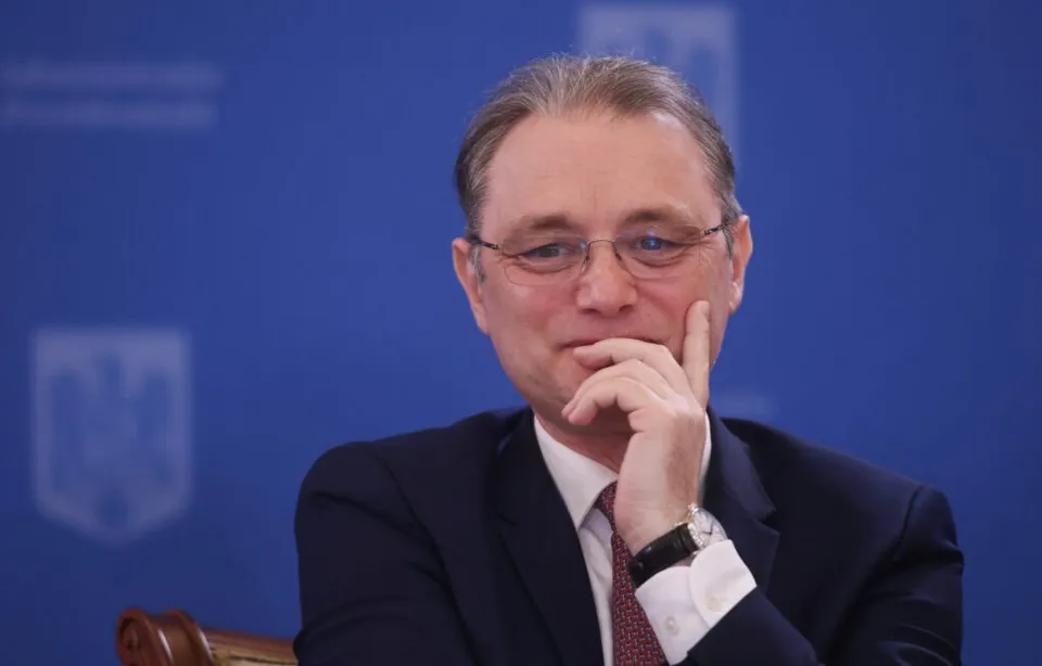 Un bărbat cu ochelari și costum închis la culoare stă cu mâna sub bărbie în fața unui fundal albastru.