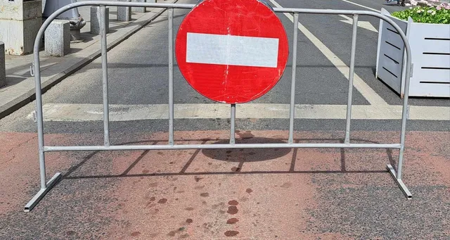 O barieră metală gri cu un semn circular roșu având o bandă albă orizontală este poziționată pe un trotuar.