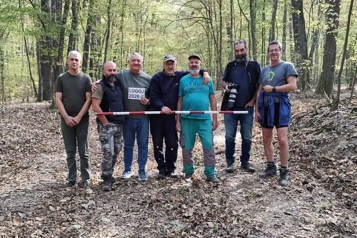 Șapte bărbați stau în rând într-o pădure, ținând împreună o bară lungă, roșie și albă.