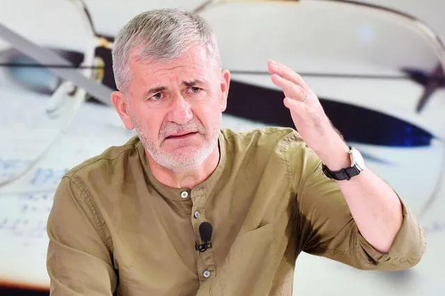 Un bărbat cu părul scurt și gri poartă o cămașă maro și ridică mâna stângă, purtând un ceas la încheietură.