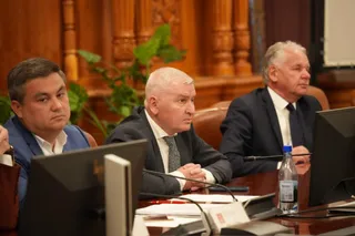 Trei bărbați în costume purtând cămași și cravate stau la o masă de conferință cu monitoare și o sticlă de apă în față.