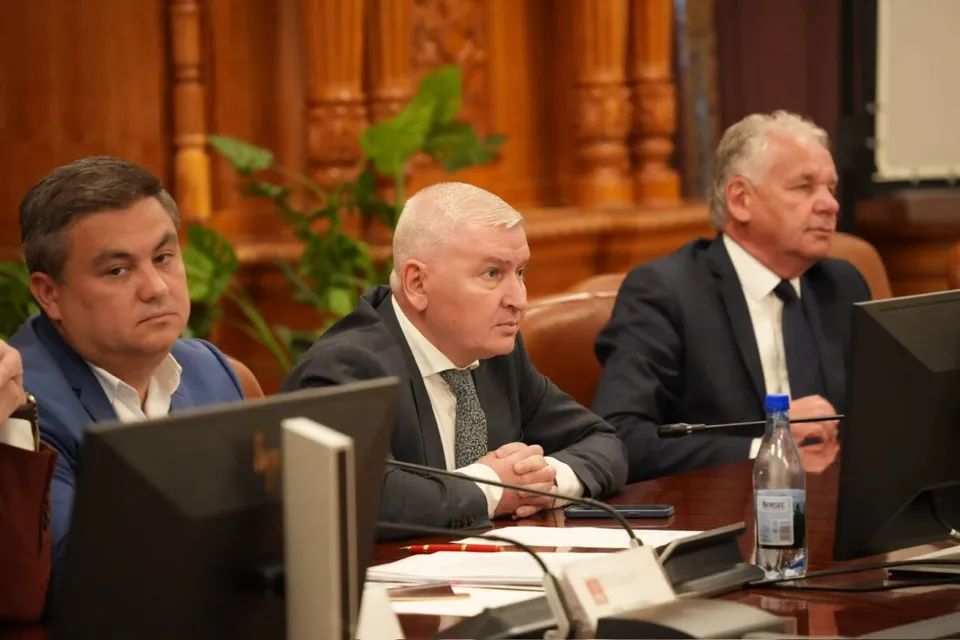 Trei bărbați în costume purtând cămași și cravate stau la o masă de conferință cu monitoare și o sticlă de apă în față.