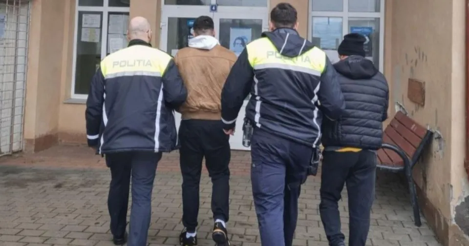 Trei polițiști în uniformă neagră și galbenă conduc doi bărbați pe un trotuar în fața unei clădiri cu ferestre mari.